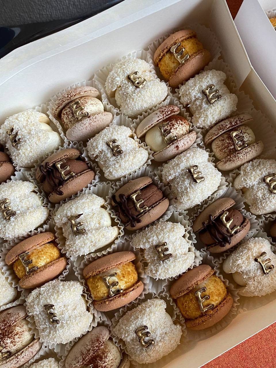 Open box of macarons in cream, blush, mocha and caramel, each with a gold chocolate letter E