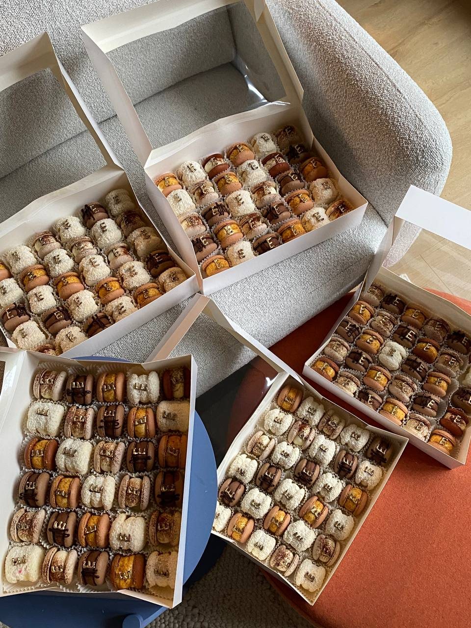 Five open white boxes of mixed macarons with gold letters, on a sofa and side tables