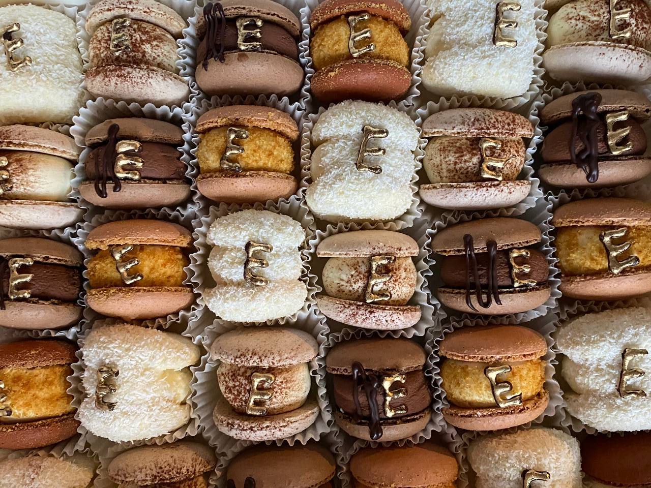 Rows of macarons seen from above: coconut-white, blush, mocha and caramel, each with a gold E