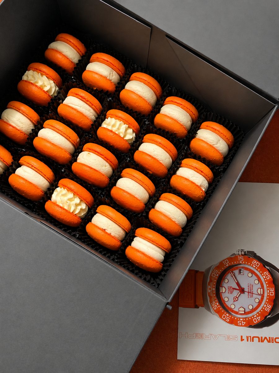 Open black gift box of 24 race-orange macarons next to a TAG Heuer Formula 1 watch card